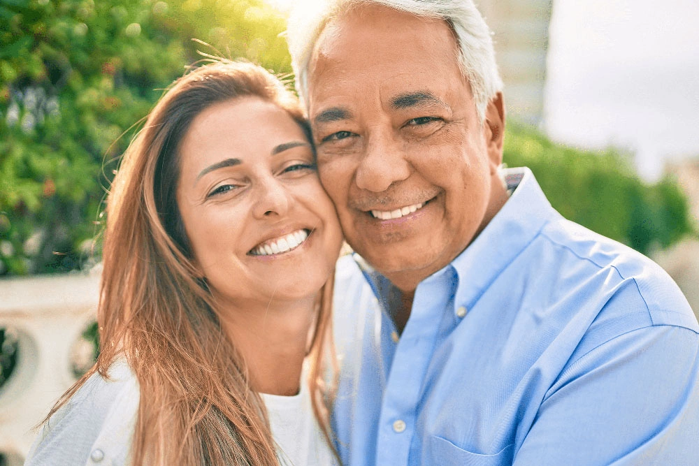 Smiling older couple cheek to cheek outdoors, illustrating smiles restored with dental bridges